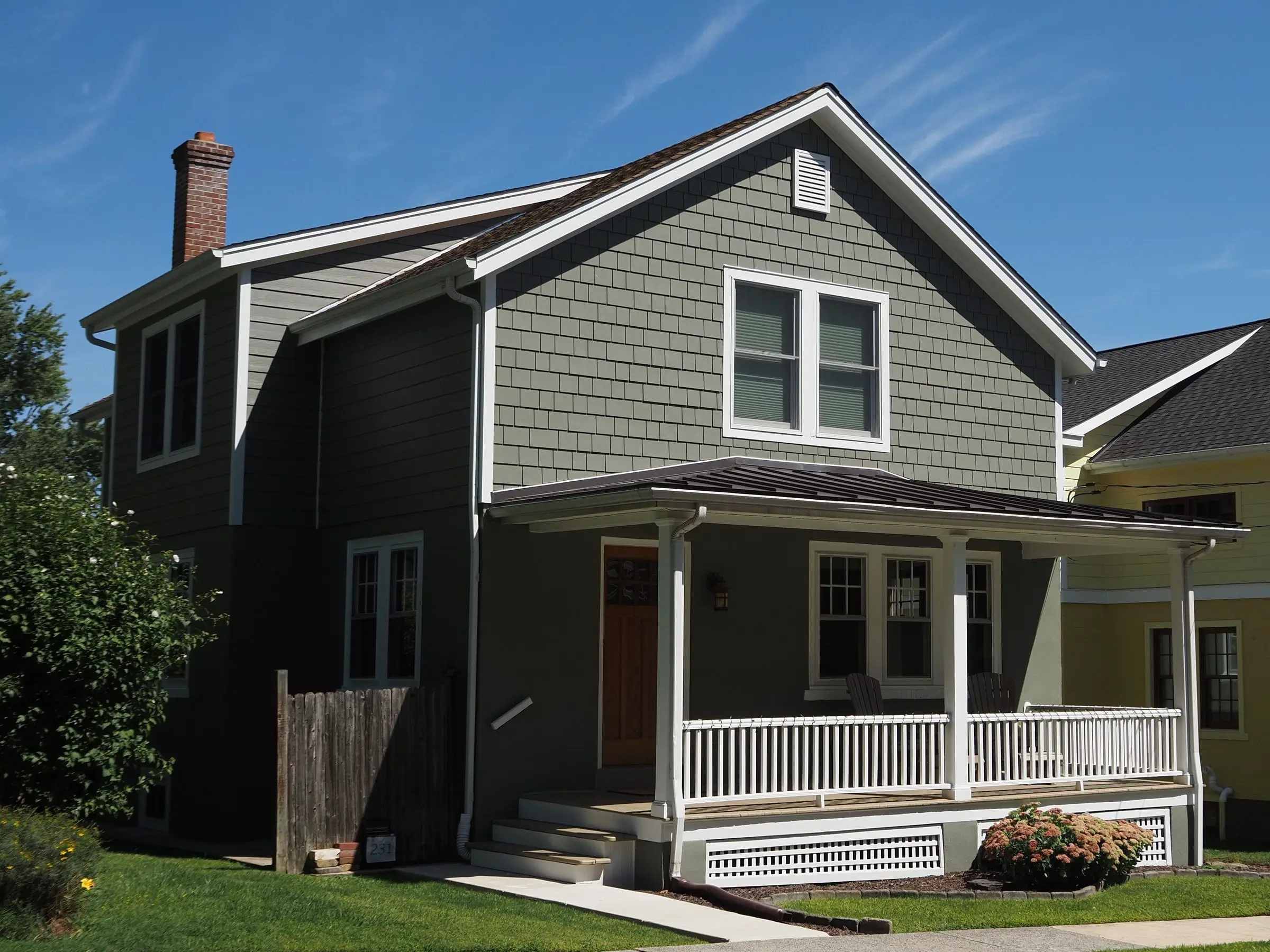 Gray shingled home with a white-railed front porch on a sunny street